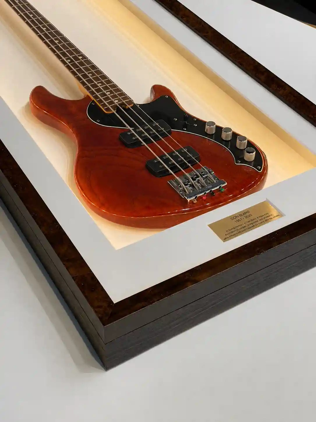 Close-up detail of a handcrafted custom shadow box frame displaying a bass guitar memorabilia presentation, featuring a polished timber veneer frame, white mount, recessed illuminated display and engraved plaque, showcasing bespoke craftsmanship.