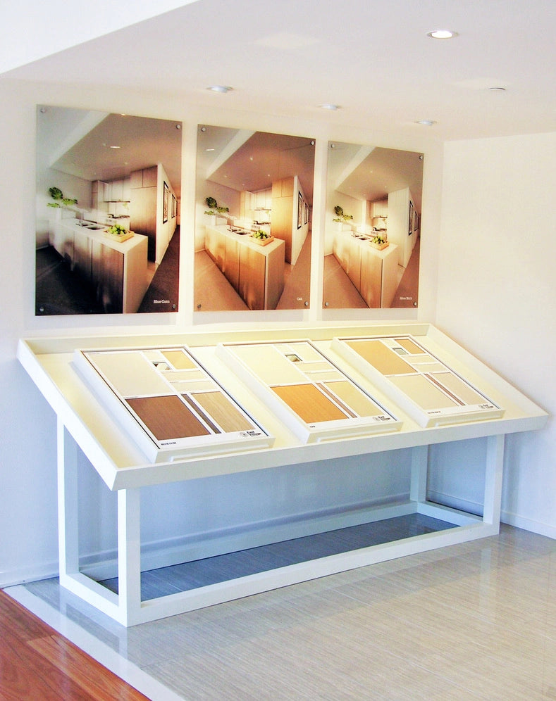 Modern showroom displaying three large kitchen finish boards above angled sample display tables featuring timber, laminate and interior finish selections in a bright contemporary space.