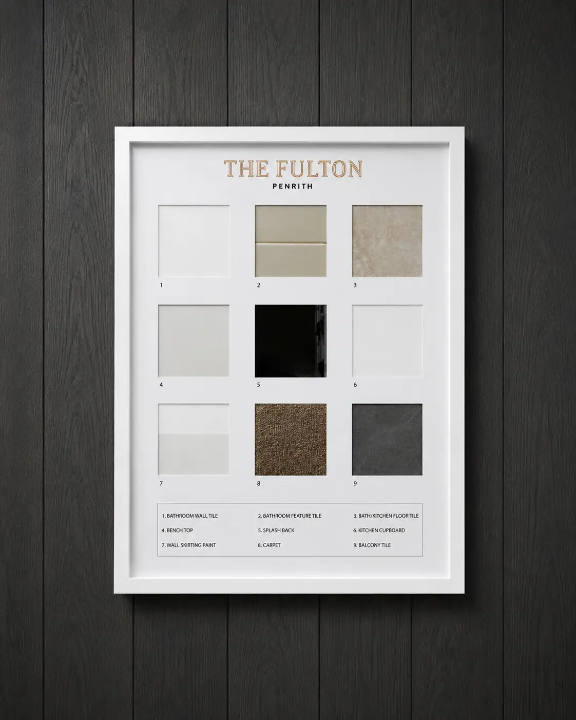White shadow box frame displaying The Fulton Penrith interior materials selection board mounted on a charcoal timber panel wall, showcasing tiles, flooring, cabinetry, paint and carpet samples in a premium presentation.
